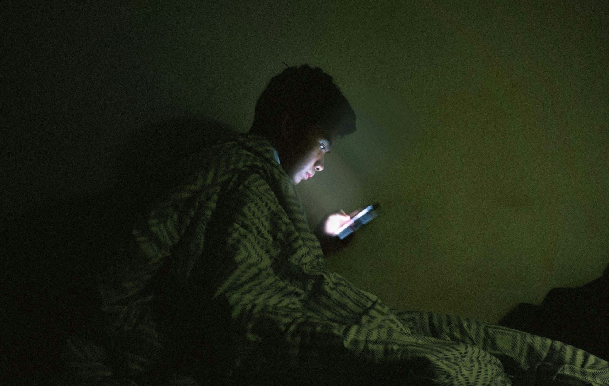 Tiener die in het donker op zijn bed zit, starend naar zijn telefoon